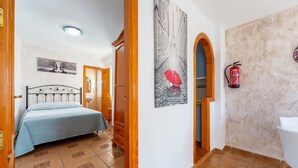A bedroom with a bed, a door leading to another room, a wall-mounted fire extinguisher, and a hallway with a tiled floor.