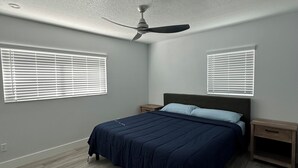 Un dormitorio con una cama, dos mesitas de noche, un ventilador de techo y dos ventanas con persianas.