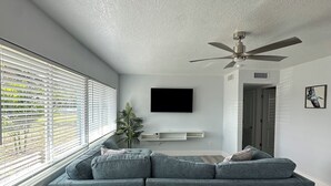 Una sala de estar moderna con un sofá seccional gris, un ventilador de techo, un televisor de pantalla plana y un gran ventanal con persianas.