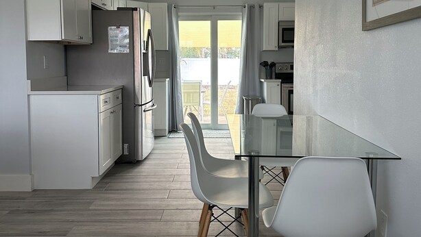 Una cocina moderna con armarios blancos, un refrigerador de acero inoxidable y un comedor con mesa de vidrio y sillas blancas.