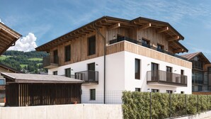 Un bâtiment moderne à plusieurs étages, doté de balcons en bois et de grandes fenêtres, entouré de verdure et surplombant un ciel dégagé.