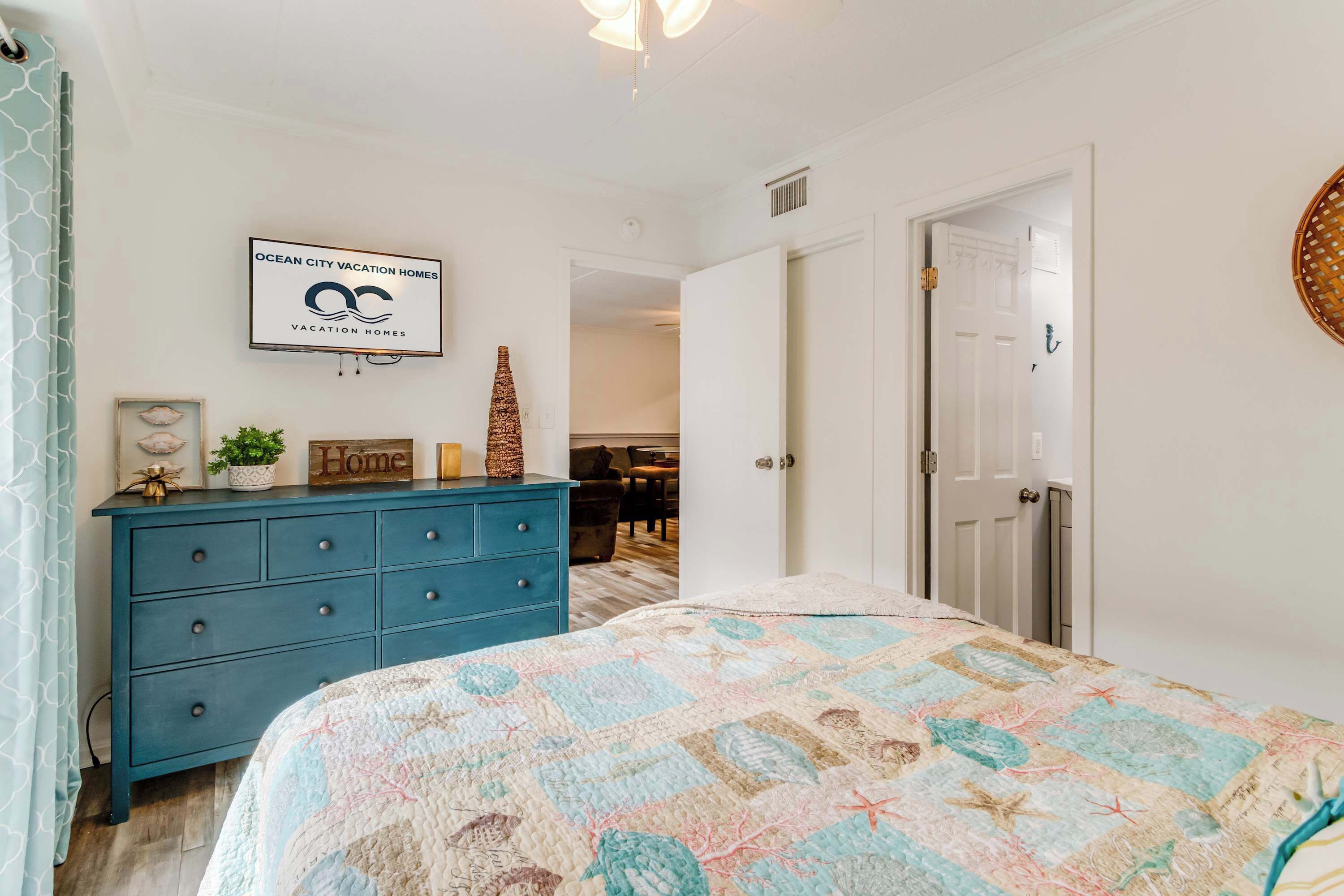 A bedroom with a blue dresser, a bed with a quilt, a framed sign on the wall, and a door leading to another room.