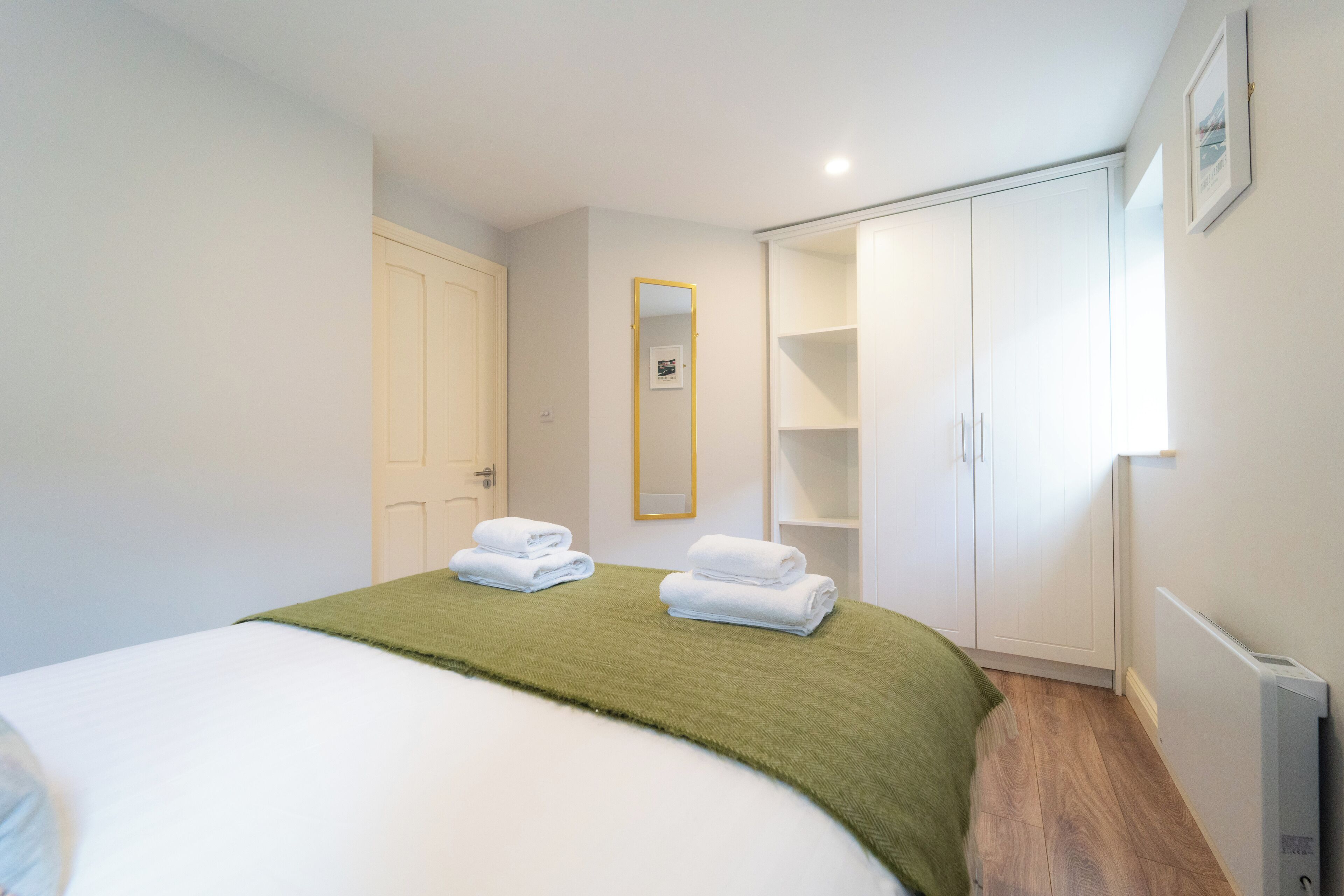 A bedroom with a bed, a green throw blanket, two neatly folded white towels, a white wardrobe, a white door, and a framed picture on the wall.