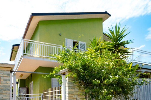 A two-story building with a balcony, a stone wall, and a tree in front.