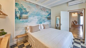 A bedroom with a large bed, a wall mural, a wooden bench, a small table, and a mirror.