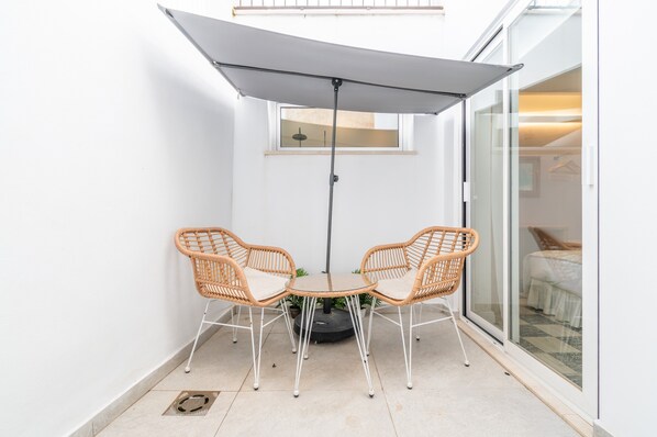 Un petit espace patio avec une table et deux chaises en osier, un grand parasol et une porte-fenêtre coulissante donnant sur une chambre.