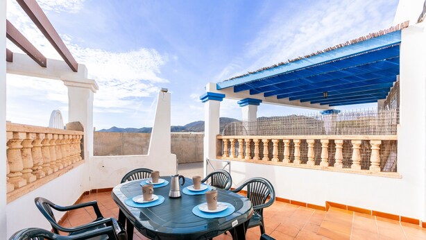 A terrace with a table set for four, chairs, a view of mountains, and a blue pergola.
