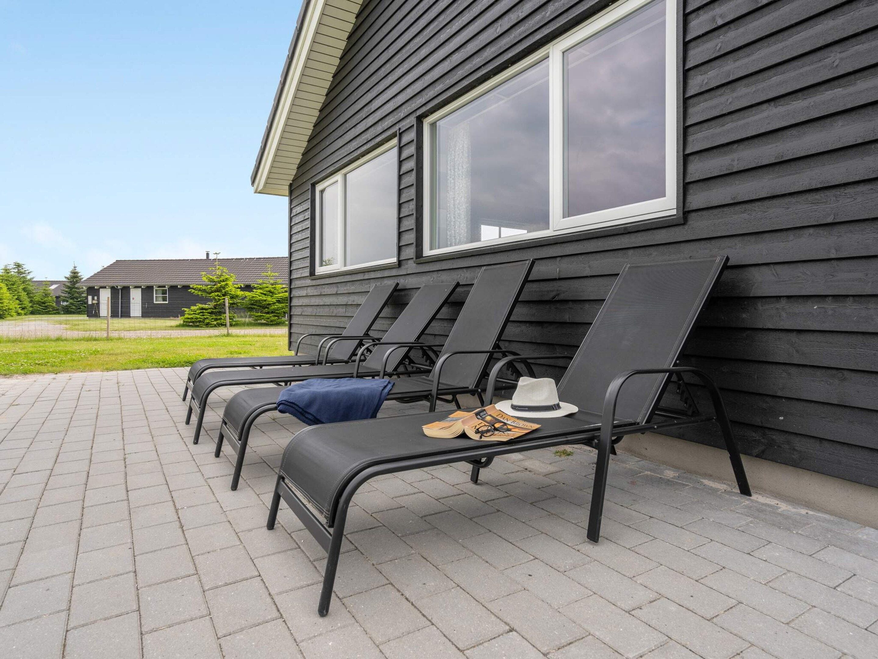 A row of outdoor lounge chairs with a hat and magazine on one, positioned in front of a building with large windows.