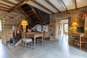 Un intérieur rustique en pierre, avec un bureau en bois, deux lampes, un escalier et un téléviseur.