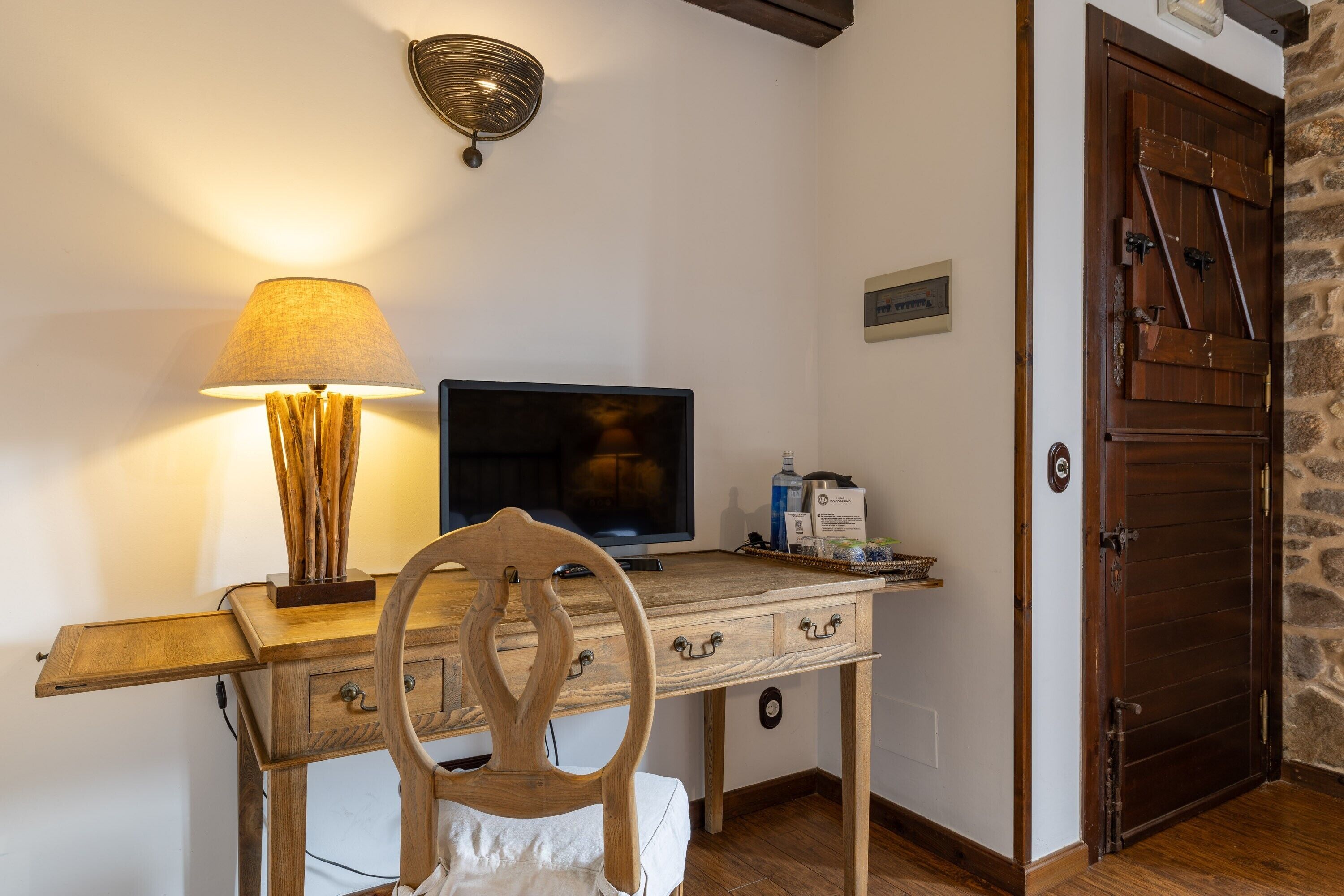 Un escritorio de madera con un televisor, una lámpara y una silla en una habitación con pared de piedra y puerta de madera.