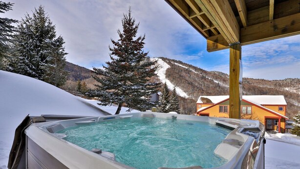 A hot tub with a mountain view, surrounded by snow-covered trees and a wooden structure. - Topridge Style 3BR/4BA with Hot Tub, Ski On Ski Off, Mountain Views and Discounted Ski Rentals (Killington)