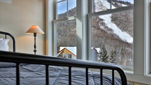 A bedroom with a bed, a lamp, and a large window with a view of a snowy mountain and a house. - Topridge Style 3BR/4BA with Hot Tub, Ski On Ski Off, Mountain Views and Discounted Ski Rentals (Killington)