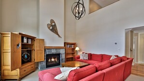 A living room with a fireplace, red sofa, wooden cabinet, and a chandelier. - Topridge Style 3BR/4BA with Hot Tub, Ski On Ski Off, Mountain Views and Discounted Ski Rentals (Killington)