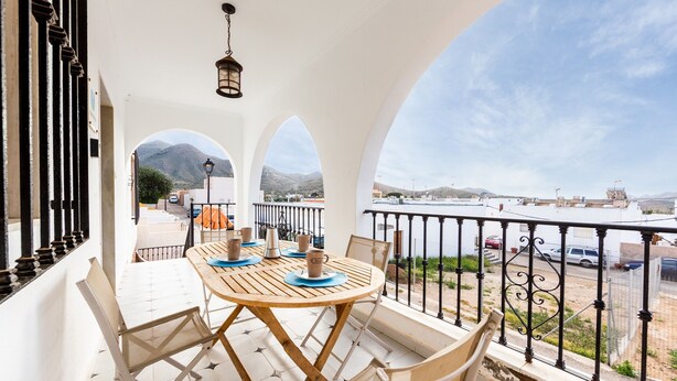 Un balcón con mesa y sillas, vista al mar y montañas al fondo.