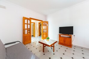 A room with a bed, a table with a plant, a TV on a wooden stand, and a door leading to another room.