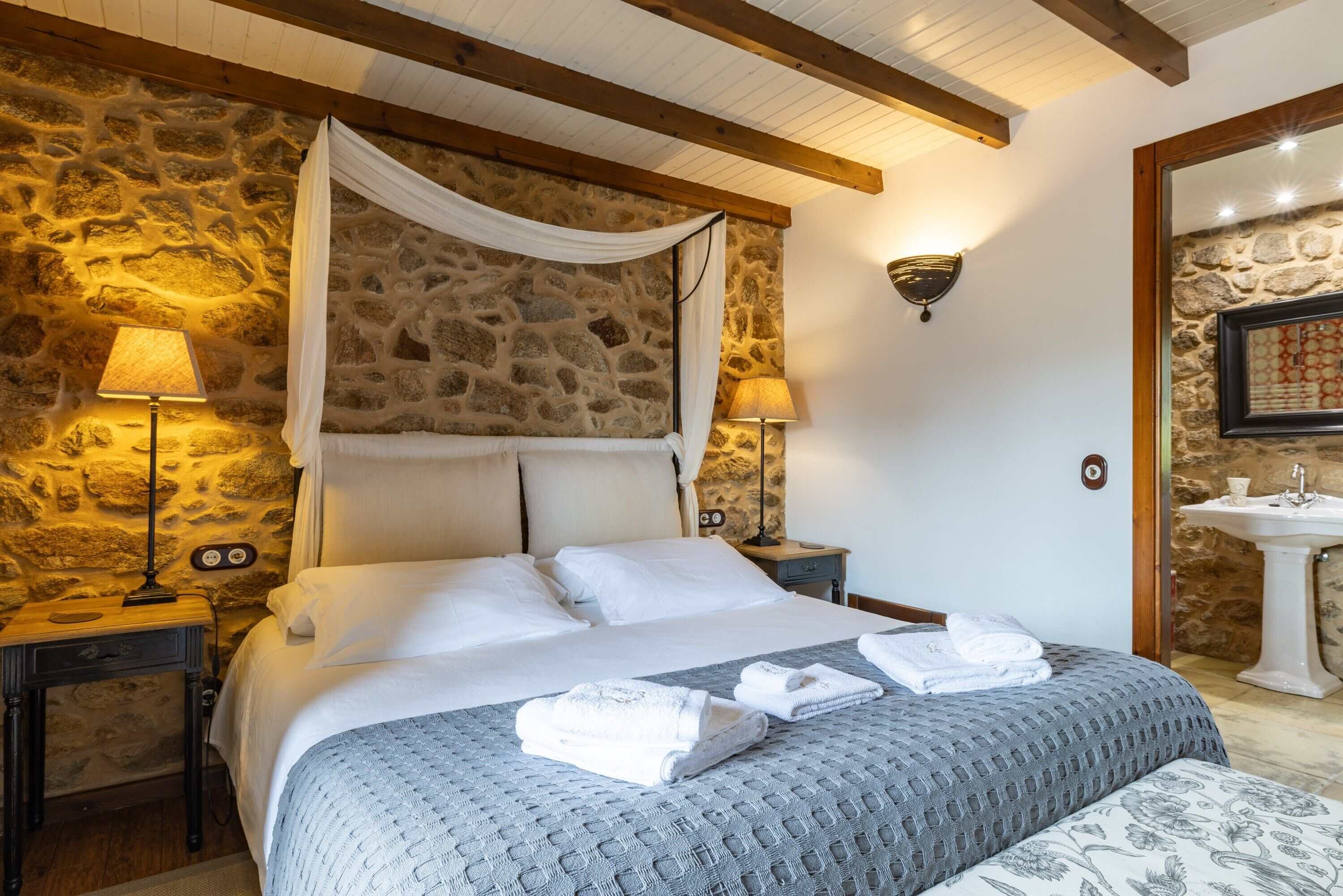 Una camera da letto con parete in pietra, un letto a baldacchino, due abat-jour, un comodino, uno specchio e un bagno con lavandino e specchio.