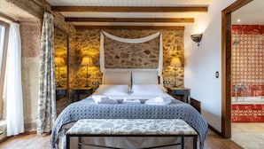 A bedroom with a stone wall, wooden beams, a bed with towels, bedside lamps, and a bathroom with patterned tiles. - Bed and Breakfast 'Alca' with Private Garden and Wi-Fi (Camariñas)