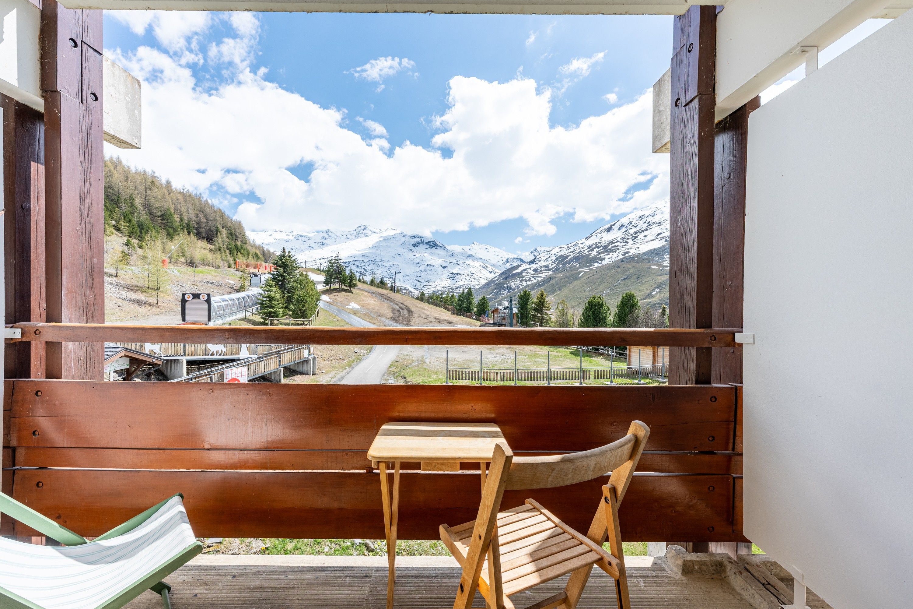 A wooden deck with a bench and a table, overlooking a mountain landscape with a road and a bridge.