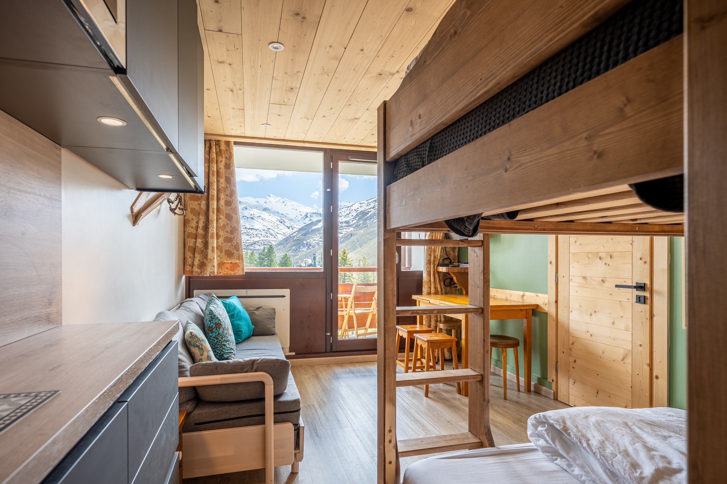 Ein kompakter, moderner Innenraum mit einer Küchenzeile, einer Couch mit Kissen, einem Essbereich mit Hocker und einem großen Fenster mit Bergblick.