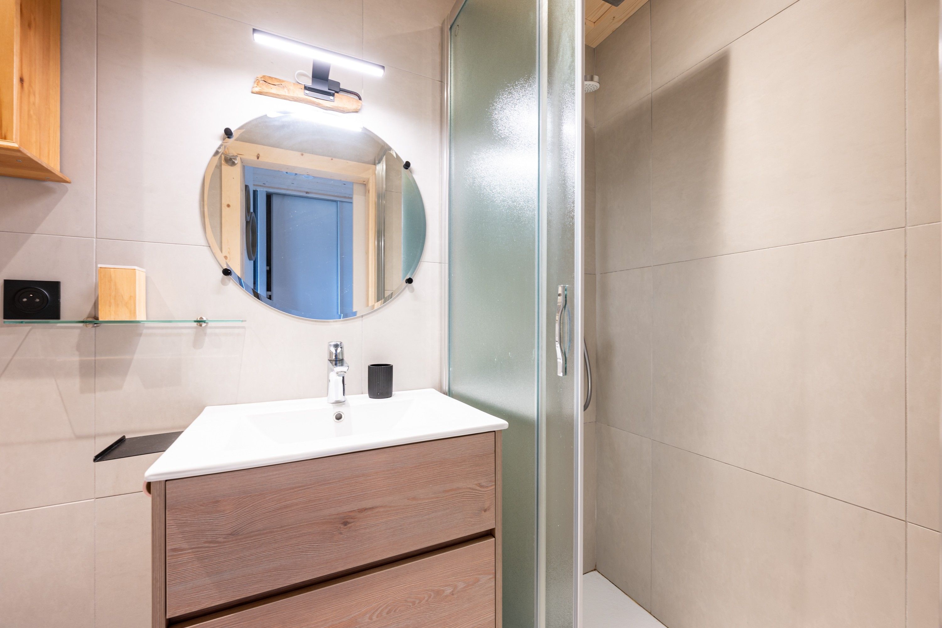 Ein Badezimmer mit einem weißen Waschbecken, einem runden Spiegel, einer Dusche mit Glastür und einem Holzkabinent.