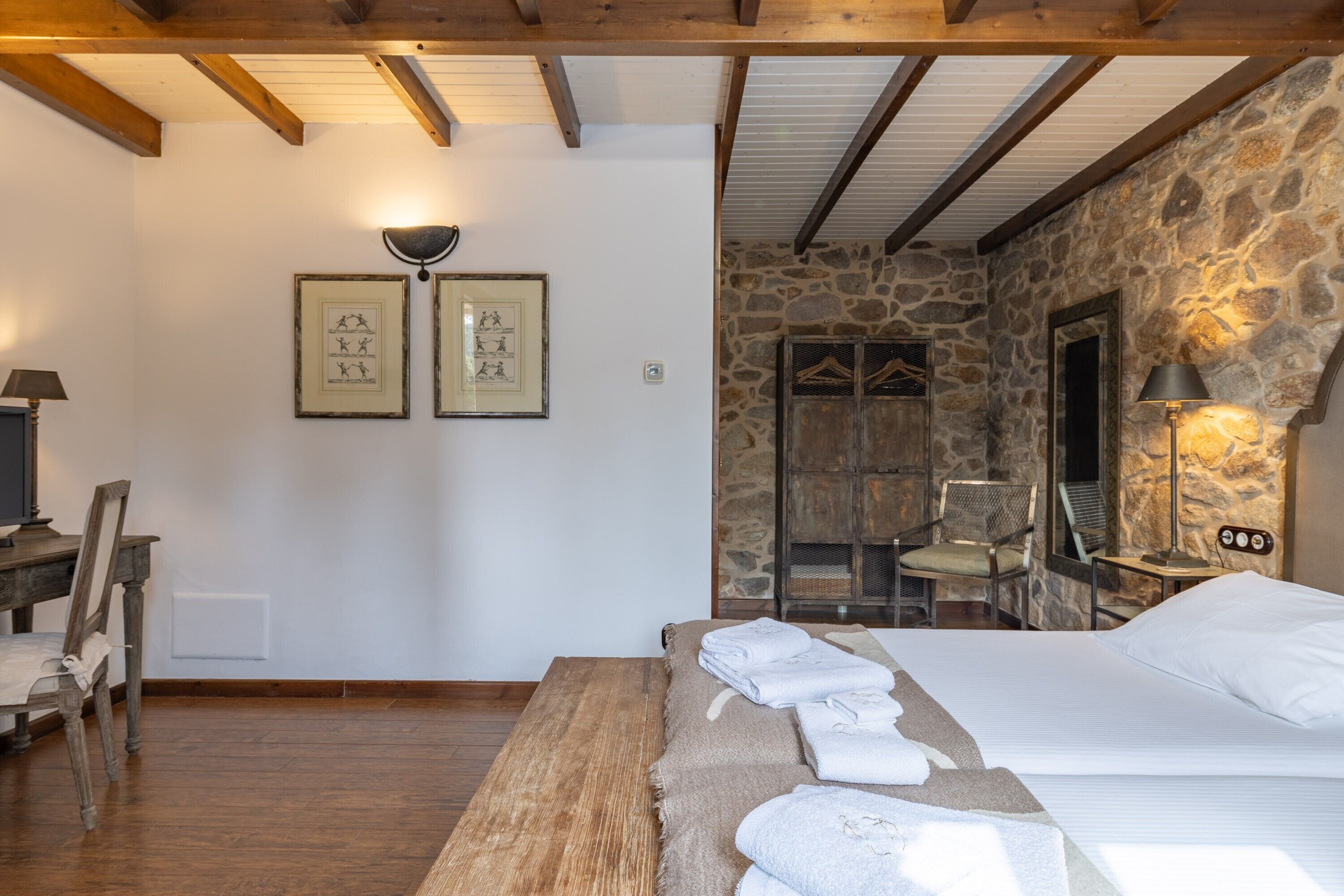 A room with a bed, a wooden table, a chair, a stone wall, and framed pictures on the wall.