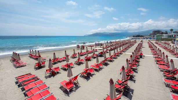 A beach with rows of red lounge chairs and umbrellas, a clear sky, and a calm sea.