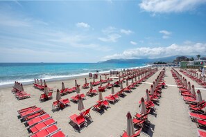 Une plage bordée de rangées de chaises longues et de parasols rouges, un ciel dégagé et une mer calme.