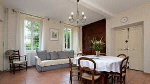 A dining area with a table set for four, a sofa with cushions, a wooden chair, a chandelier, and a view of greenery outside. - Maison L'Arénoulat in Aspet – charm, garden & comfort in the heart of Comminges, near town & nature (Aspet)