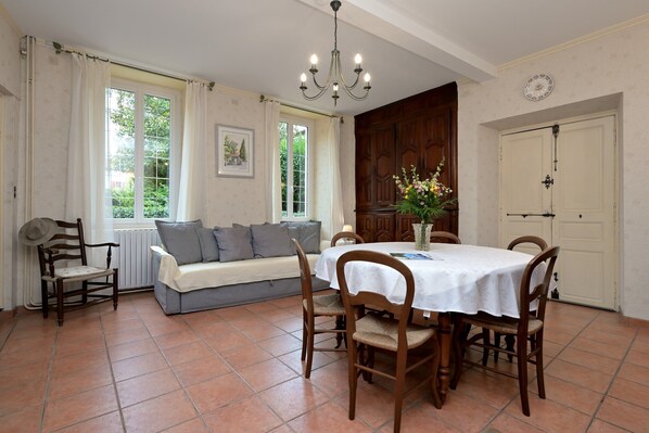 A dining area with a table set for four, a sofa with cushions, a wooden chair, a chandelier, and a view of greenery outside.