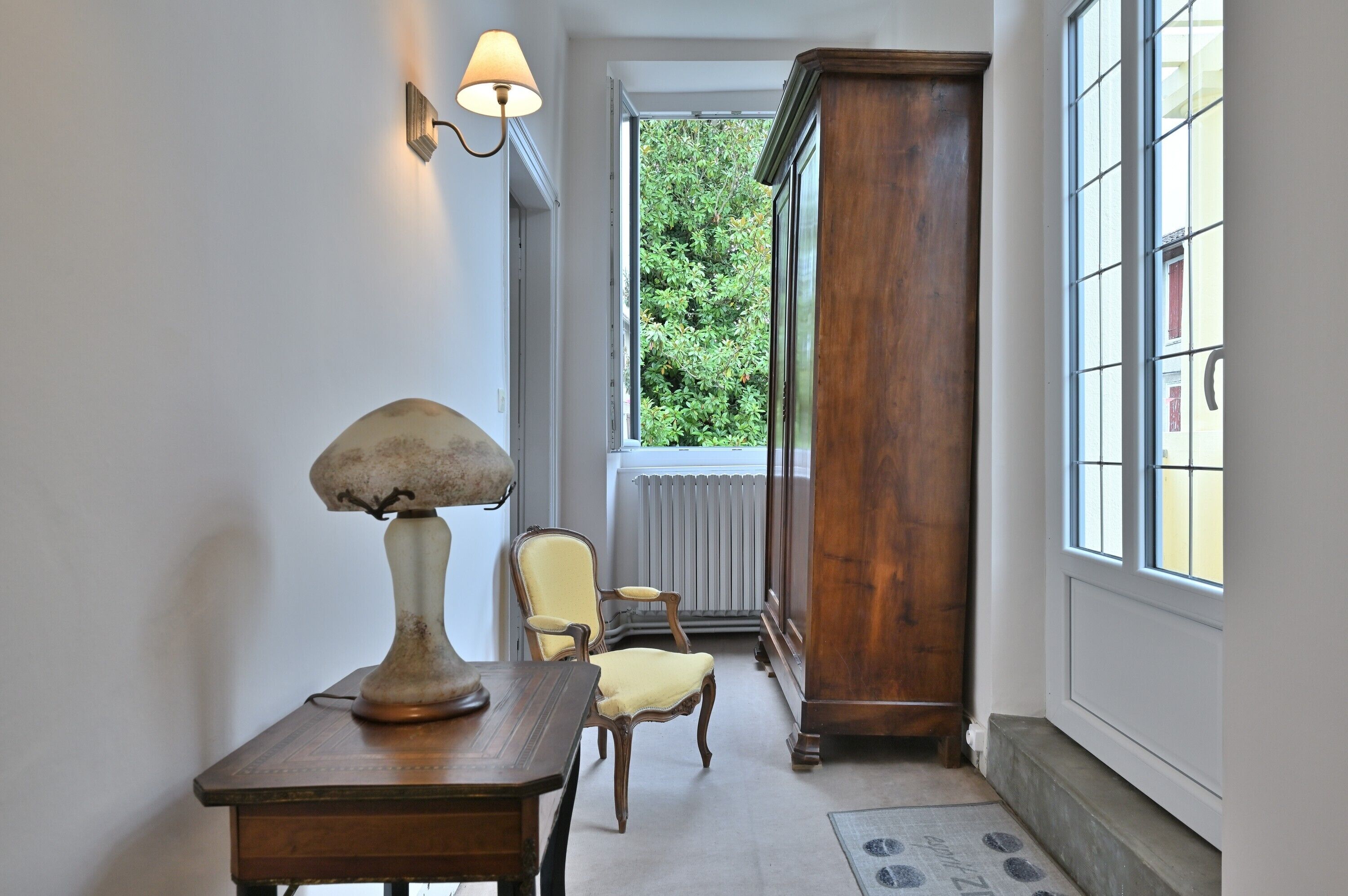 A hallway with a lamp, chair, wooden cabinet, and a window with a view of greenery.