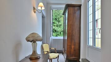 A hallway with a lamp, chair, wooden cabinet, and a window with a view of greenery.
