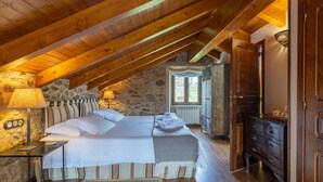 A bedroom with a stone wall, wooden ceiling beams, a bed with white linens, a bedside table with a lamp, and a dresser.
