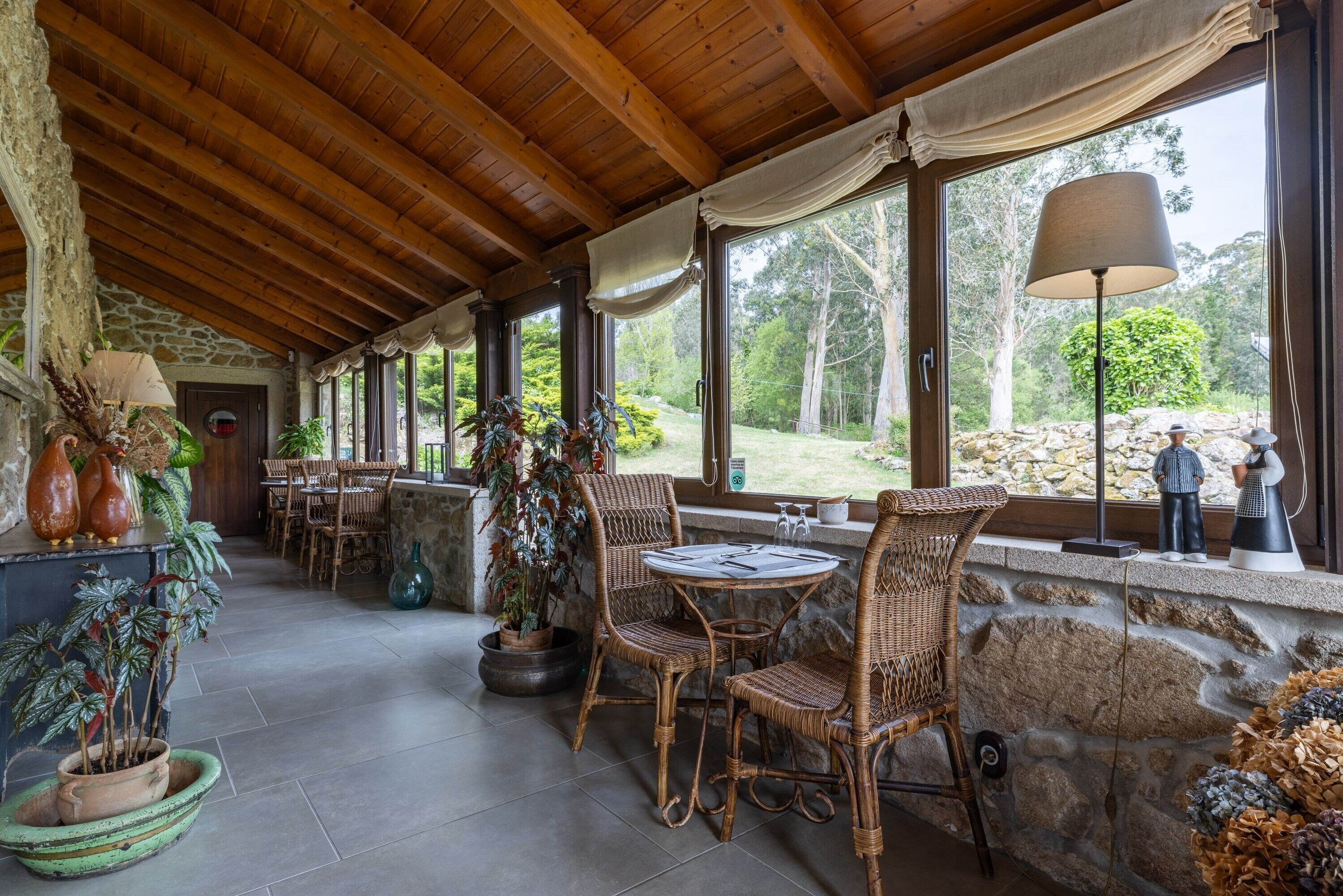 A covered patio area with wicker furniture, stone walls, and large windows overlooking a natural landscape.