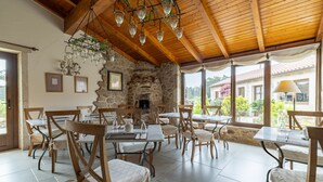 Un comedor con paredes de piedra, vigas de madera en el techo y lámparas colgantes. Mesas y sillas dispuestas para comer.