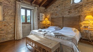 Un dormitorio con paredes de piedra y techo de madera, una cama con ropa blanca, un banco y una mesita de noche con lámparas.