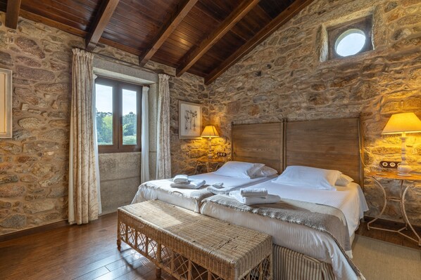 Un dormitorio con paredes de piedra y techo de madera, una cama con ropa blanca, un banco y una mesita de noche con lámparas.