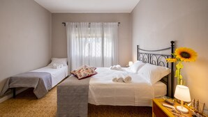 Une chambre d’hôtel avec deux lits, une table de chevet avec une lampe, un vase contenant un tournesol et une fenêtre avec des rideaux légers.