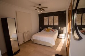 Ein Schlafzimmer mit einem Bett, einem Deckenventilator, einem Heizkörper, einem Nachttisch mit Lampe und einem großen Spiegel.