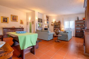 A living room with a sofa, a dining table with a green tablecloth, a television, and a bookshelf. - Country House 'La Villa Del Relax' with view on the rolling hills, Terrace and Wi-Fi (Sennori)