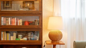 Ein Bücherregal aus Holz mit Büchern und dekorativen Gegenständen, eine Korbtasche mit einer Lampe auf einem kleinen Tisch und ein Stuhl.