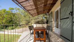 Eine überdachte Terrasse mit Holztisch und -stühlen, einem Metallzaun und Blick auf Bäume und ein Gebäude.