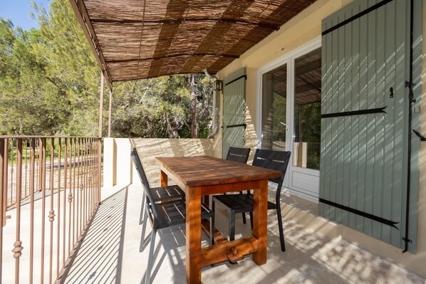 A covered outdoor patio with a wooden table and chairs, a metal railing, and a view of trees and a building in the background.