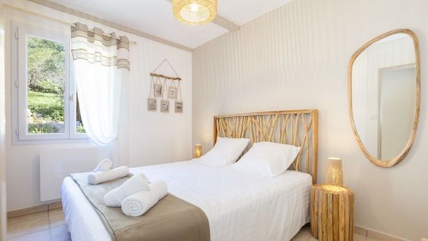 Una cama bien hecha con ropa de cama blanca y un detalle en tonos marrones, una lámpara de noche, un espejo y una ventana con cortinas.