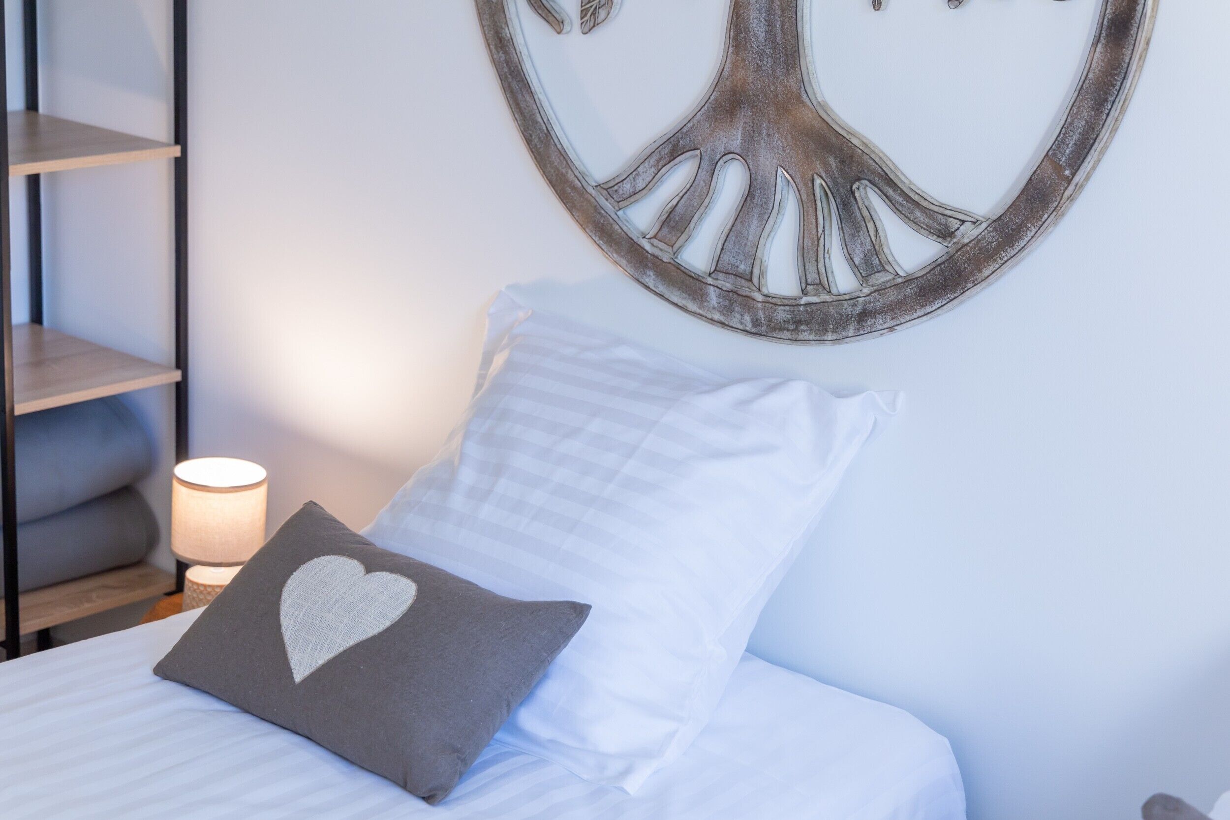 A neatly made bed with a pillow featuring a heart design, a wall-mounted tree decoration, and a bedside lamp.