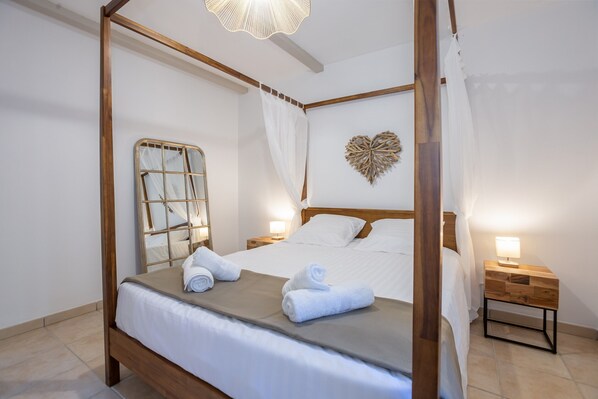 Ein Bett mit Baldachin, weiße Bettwäsche, ein Nachttisch mit Lampe und ein dekoratives Herz an der Wand.