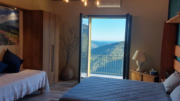 Un dormitorio con una cama grande, una cama más pequeña, una mesita de noche con una lámpara, un florero con ramas y un ventanal con vistas a la montaña.