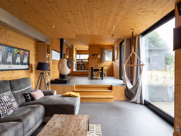 Ein modernes Interieur mit Holzbalkendecke, einer Couch, einem Tisch, einem Hängekorbstuhl und Blick durch große Fenster.