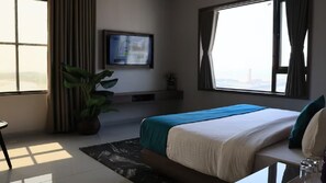 Ein Hotelzimmer mit Bett, Fernseher an der Wand, großem Fenster und einer Zimmerpflanze.