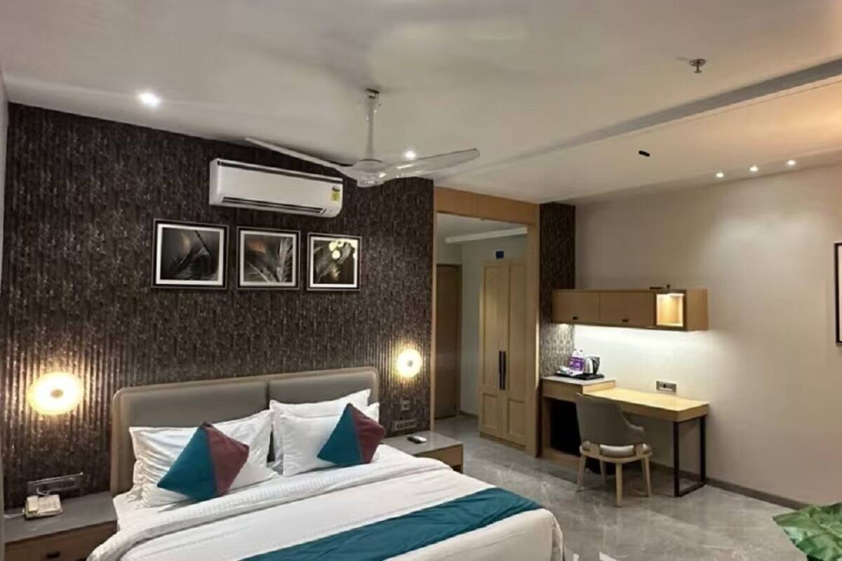 A modern hotel room with a bed, desk, and chair, featuring a dark textured wall, ceiling fan, and artwork.