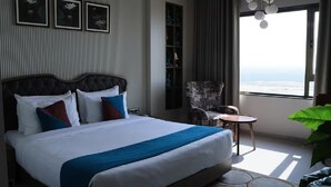 A hotel room with a large bed, a small round table, a chair, a bookshelf, and a large window with a view. - Pride Elite Dwarka (Dwarka)